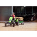 child-driving-a-green-berg-pedal-tractor-with-a-hay-loaded-trailer-outside-a-cow-barn