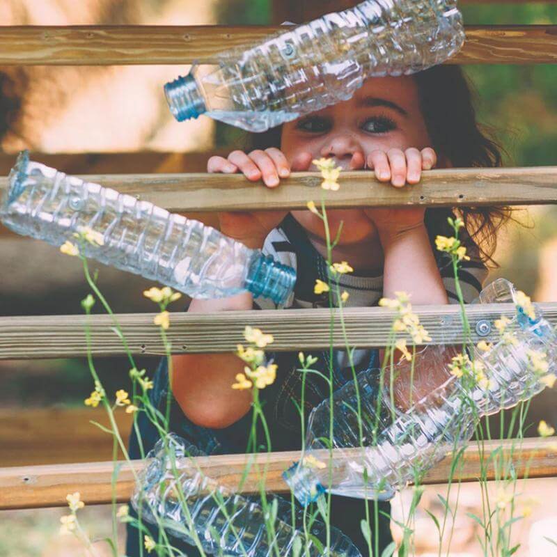 child-peeking-through-wooden-playcentre-wall-with-hanging-plastic-bottles