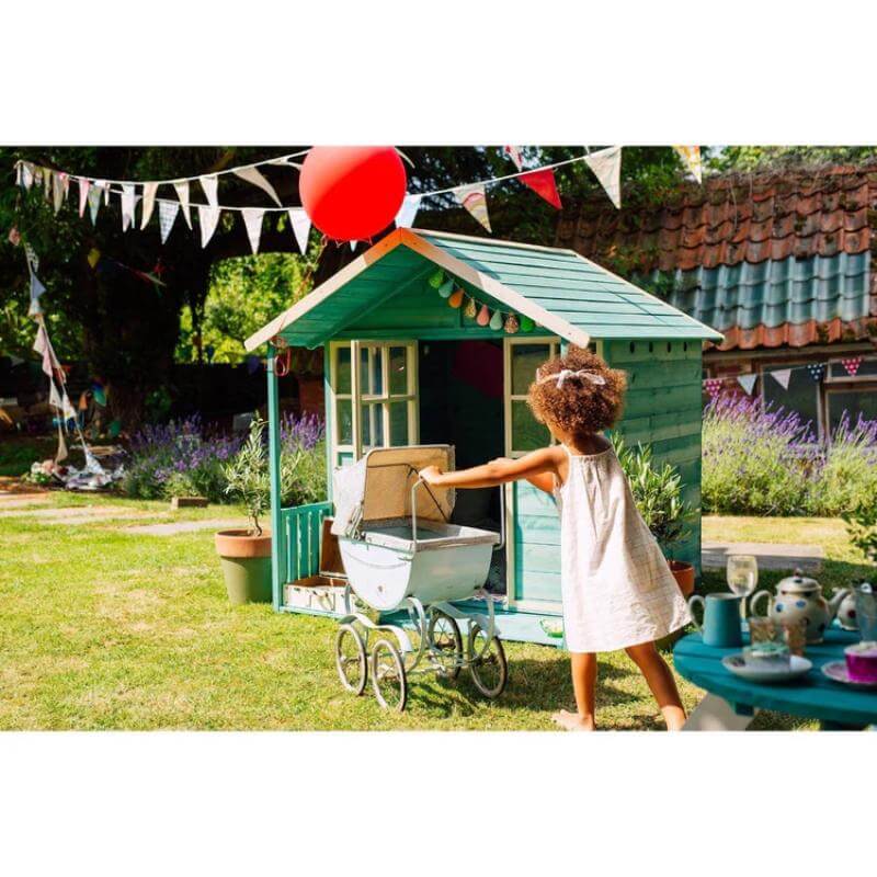 child-pushing-vintage-toy-pram-towards-teal-wooden-playhouse-in-garden