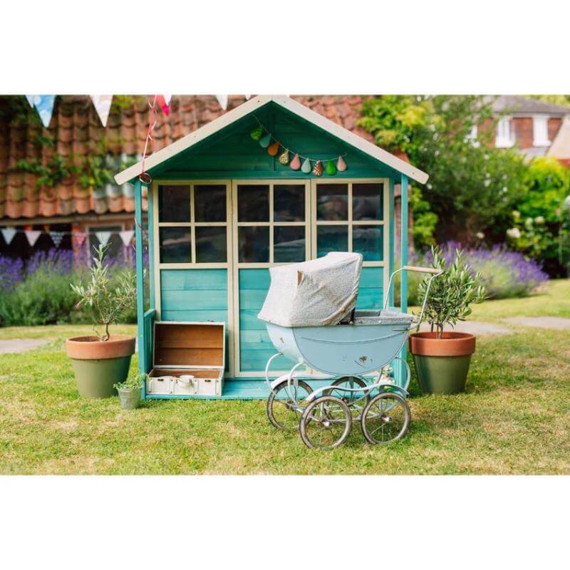front-view-of-teal-wooden-playhouse-in-garden-with-toy-pram-and-potted-plants