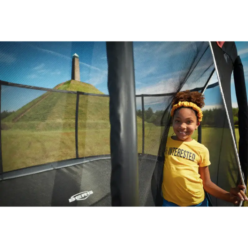 girl-standing-at-berg-trampoline-safety-net-entrance-with-grassy-hill-in-background
