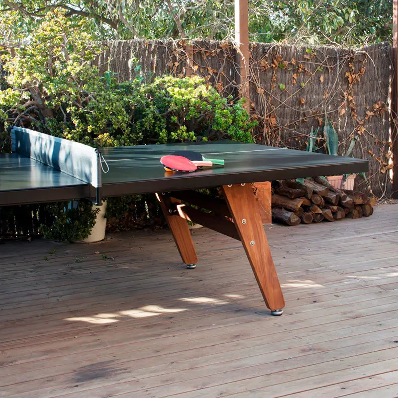 outdoor-barcelona-stationary-ping-pong-table-with-net-and-tabletop-and-legs-detail-with-paddles-on-the-surface-and-plants-and-firewood-logs-in-the-background